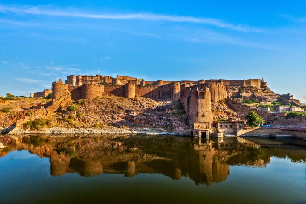 Mehrangarh Fort in Jodhpur, India, reflected in the still waters of a surrounding lake, with golden sandstone walls glowing under clear blue skies.