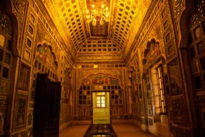 Ornately decorated interior hall of Mehrangarh Fort featuring intricate mirror work, colorful frescoes, and a glowing chandelier under a vaulted ceiling.