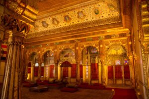 The royal Durbar Hall inside Mehrangarh Fort, richly adorned with golden arches, intricate frescoes, stained glass windows, and a regal throne canopy.
