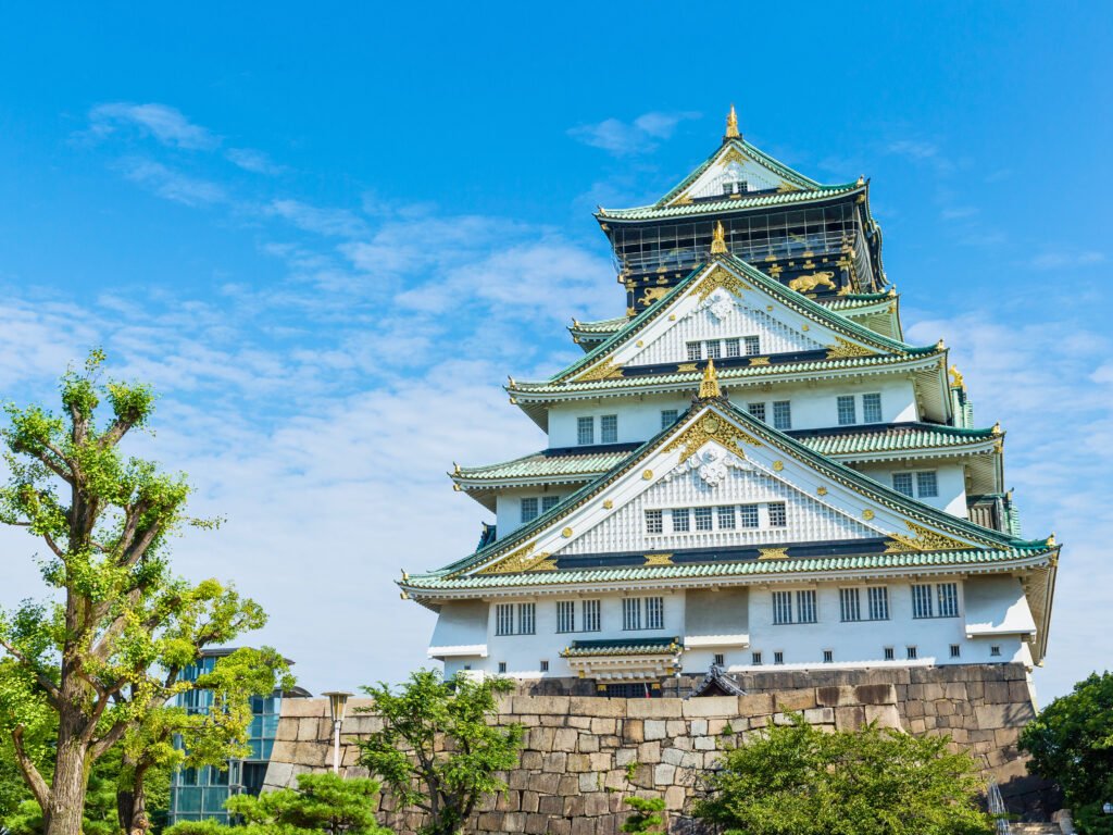 Front view of Osaka Castle in Japan, showcasing its traditional white walls, green-tiled roofs, and golden accents under a bright blue sky.