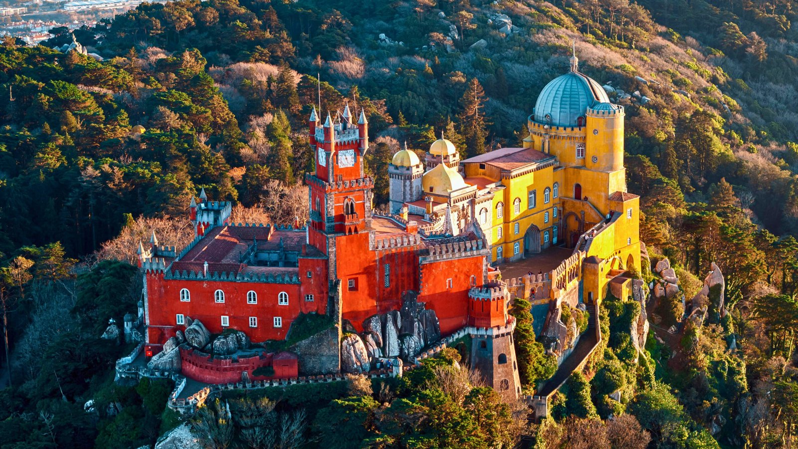 Aerial view of the colorful Pena Palace in Sintra, Portugal, showcasing its vibrant red and yellow towers surrounded by lush forested hills.