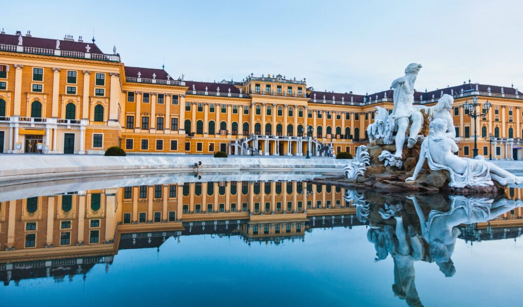 Reflection of Schönbrunn Palace in a still fountain pool with classical statues in the foreground, capturing the Baroque architecture at twilight.