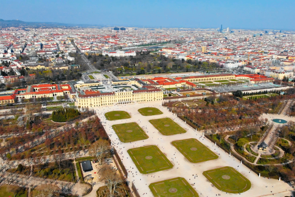 Aerial view of Schönbrunn Palace and its symmetrical Baroque gardens, surrounded by the cityscape of Vienna on a clear day.