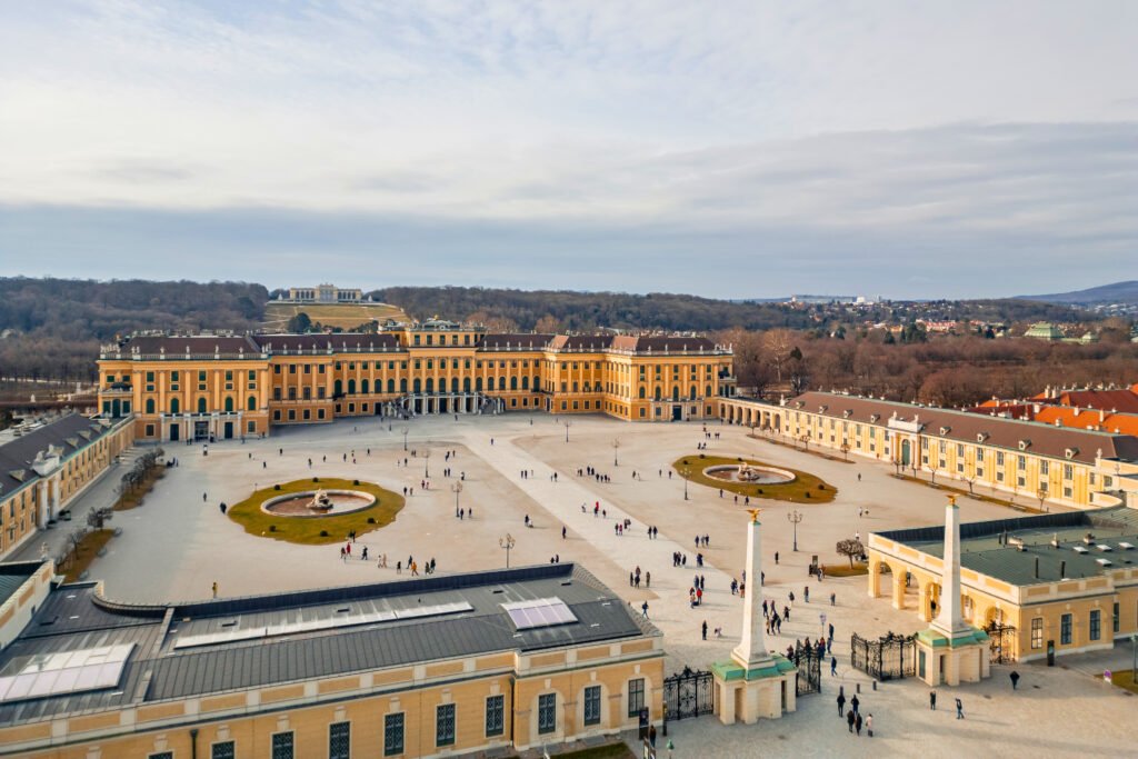 Aerial view of the grand forecourt of Schönbrunn Palace, with symmetrical layout, fountains, and distant hills framing the Gloriette in the background.