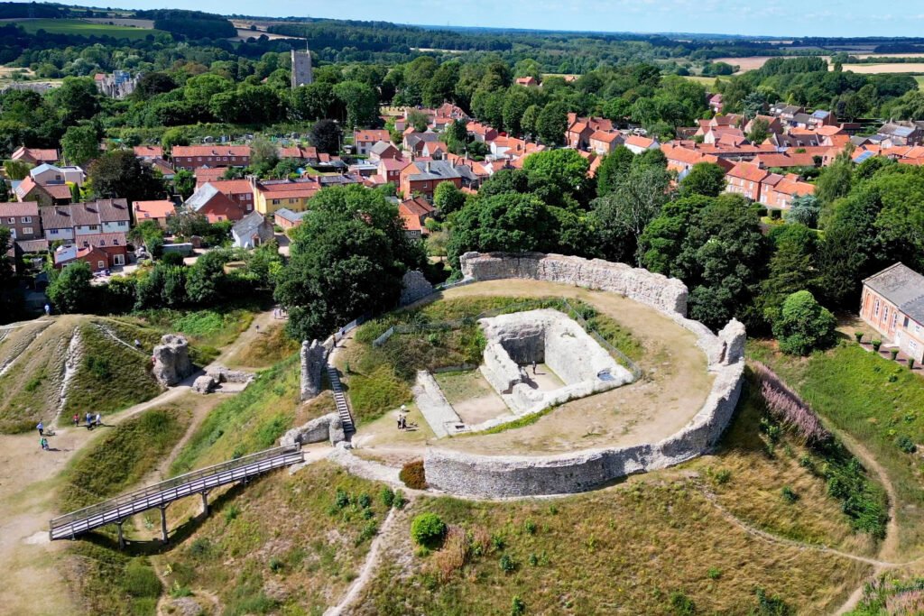 Aerial view of Castle Acre Castle in Norfolk, England, showing the motte, stone keep ruins, surrounding ditch, and wooden footbridge.