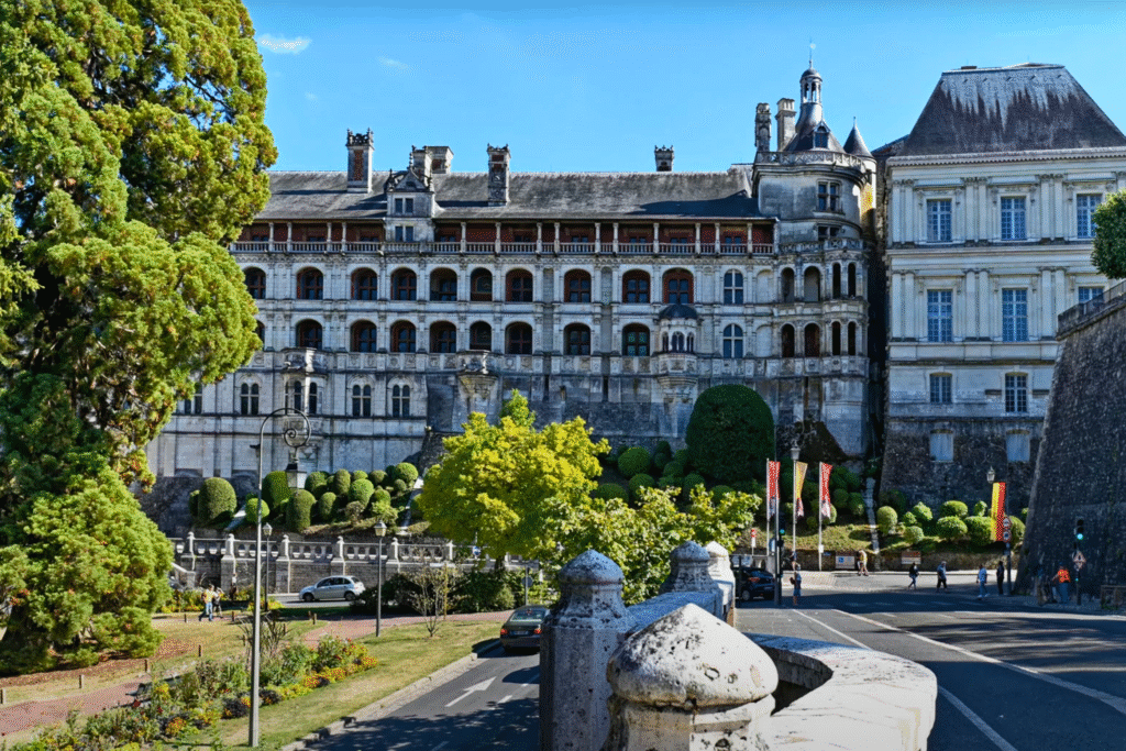 Renaissance façade of Château de Blois seen from the street, framed by gardens, trees, and historic buildings under a clear blue sky