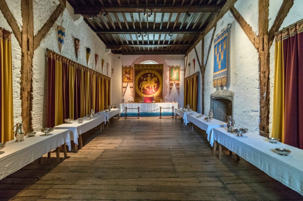 Interior of a medieval castle great hall with long banquet tables, heraldic banners, and timber-framed walls.