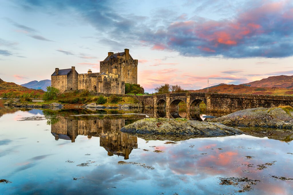 Eilean Donan Castle at sunrise reflected in still water, surrounded by Scottish Highlands and dramatic skies.