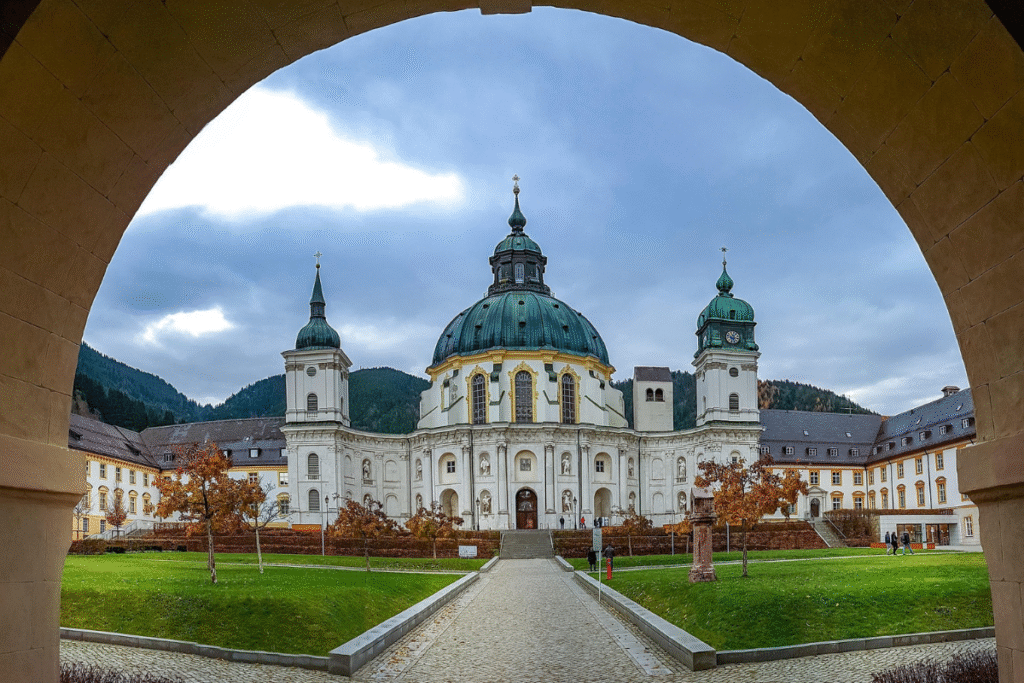 Frontal view of Ettal Abbey’s Baroque facade and dome, framed by an archway with the Bavarian Alps in the background.