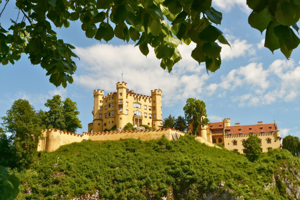 View of Hohenschwangau Castle on a sunny day, framed by tree leaves, with the bright yellow facade rising above green hills in Bavaria, Germany.