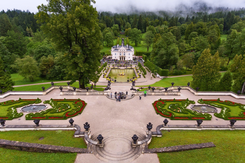 Aerial view of Linderhof Palace framed by formal gardens, fountains, and forested Bavarian hills.