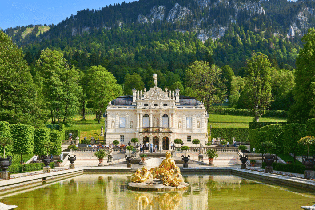 Front view of Linderhof Palace with its golden fountain and manicured gardens, set against a forested Bavarian hillside.