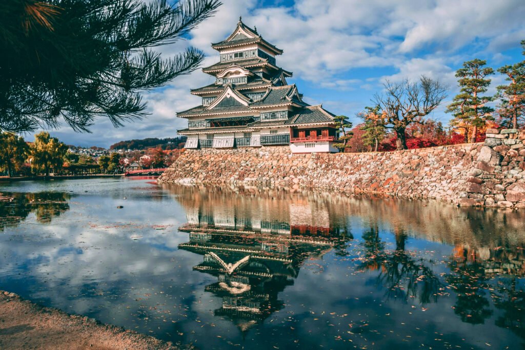 Matsumoto Castle reflected in its moat during autumn, with colorful foliage and stone walls enhancing the tranquil scenery in Nagano, Japan.