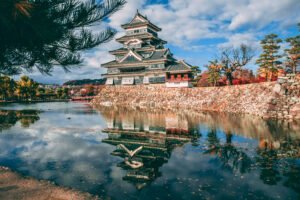 Matsumoto Castle reflected in its moat under a blue sky, surrounded by fall foliage.
