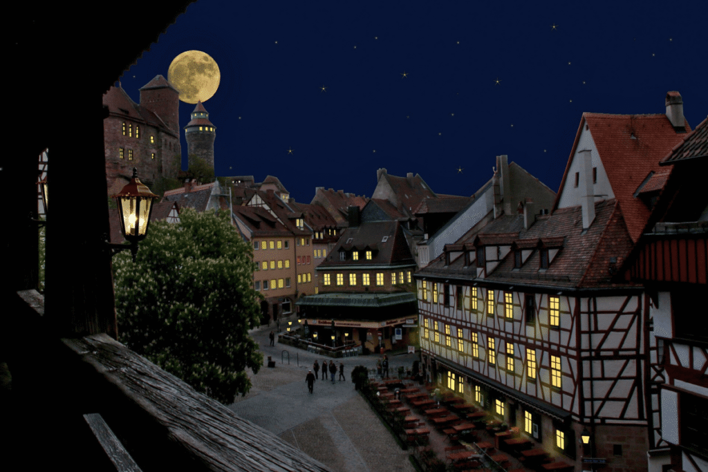 Night view of Nuremberg Castle and Old Town under a starry sky, with illuminated timber-frame houses and a full moon rising above Kaiserburg Nürnberg.
