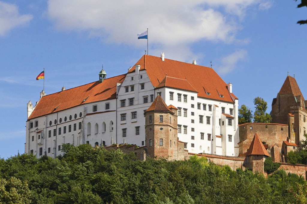 View of Trausnitz Castle in Landshut, Germany, with its white walls and red-tiled roofs perched above green trees on a sunny day.