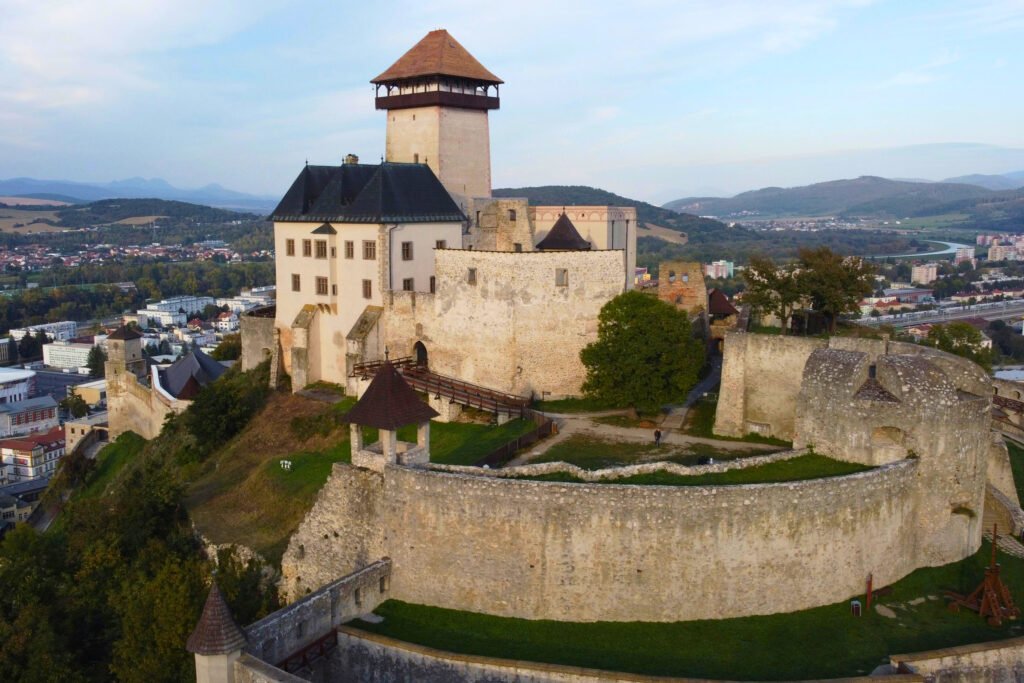Aerial view of Trencin Castle in Slovakia, perched on a rocky hilltop above the town with stone ramparts, a central tower, and panoramic views of the surrounding landscape.