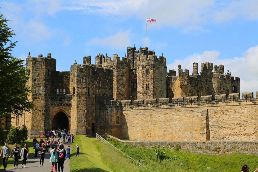 Visitors walk toward the entrance of Alnwick Castle, its stone towers and battlements flying the Union Jack under a blue sky.