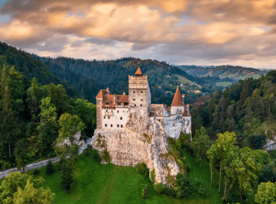 Aerial view of Bran Castle at sunset, perched on a rocky hill and surrounded by forest in Transylvania, Romania.