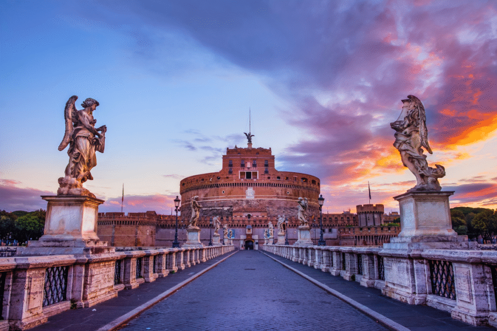 Castel Sant’Angelo in Rome at sunrise, viewed from Ponte Sant’Angelo with angel statues and a dramatic sky overhead.