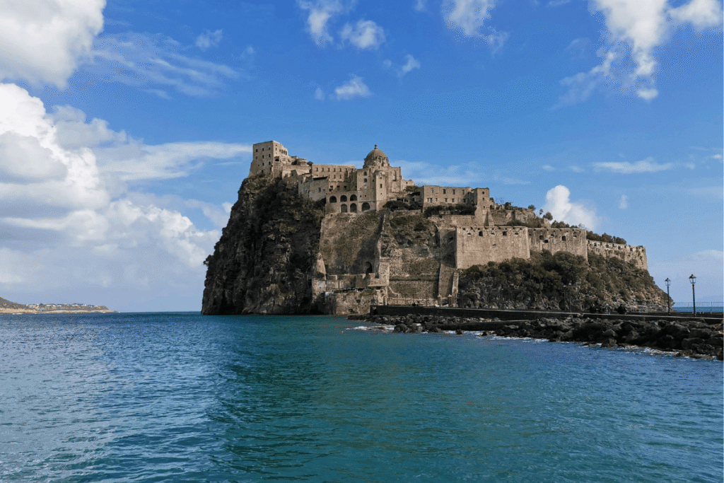 Castello Aragonese on Ischia, Italy, perched on a rocky islet above the sea under a blue sky with scattered clouds.