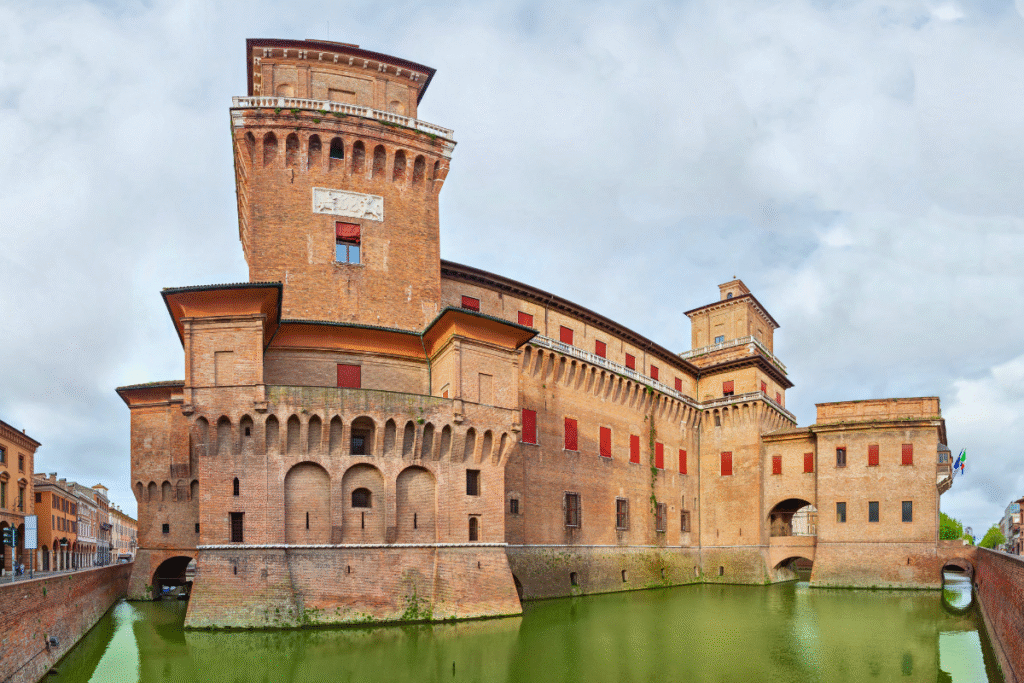 Castello Estense in Ferrara, Italy, surrounded by its original moat and featuring brick towers with red shutters under a cloudy sky.