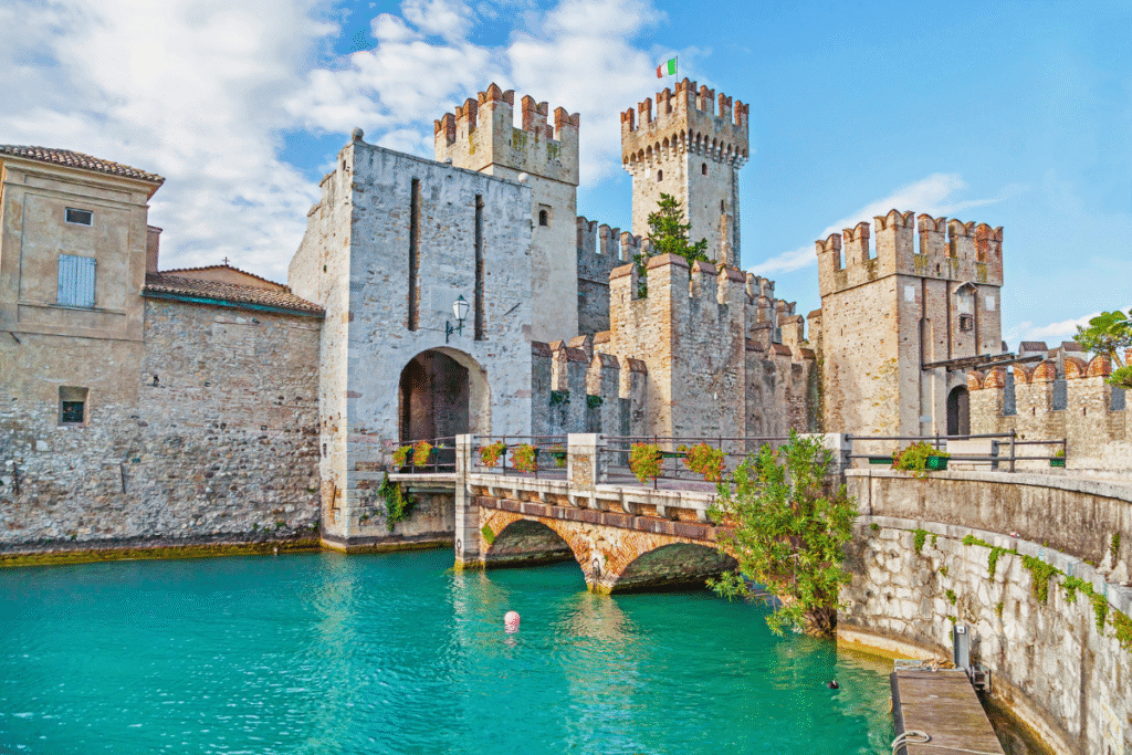 View of Castello Scaligero in Sirmione, Italy, with its medieval stone towers and moat filled with turquoise lake water under a bright blue sky.