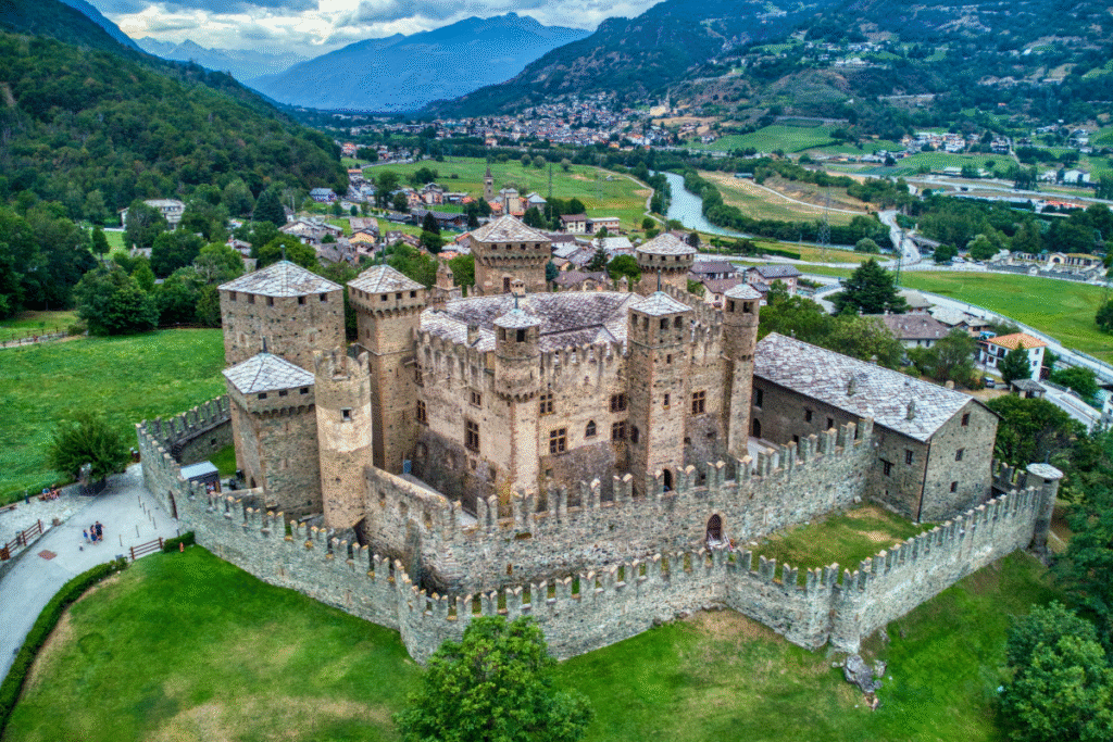 Castello di Fenis in the Aosta Valley, Italy, seen from above with its many towers and stone walls surrounded by green fields and mountains.