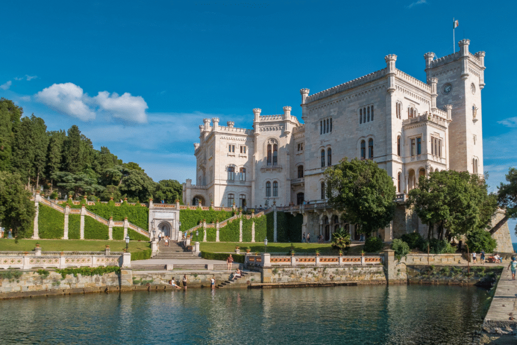 Castello di Miramare in Trieste, Italy, viewed from the water with its white neo-Gothic façade, grand staircase, and lush gardens on a sunny day.