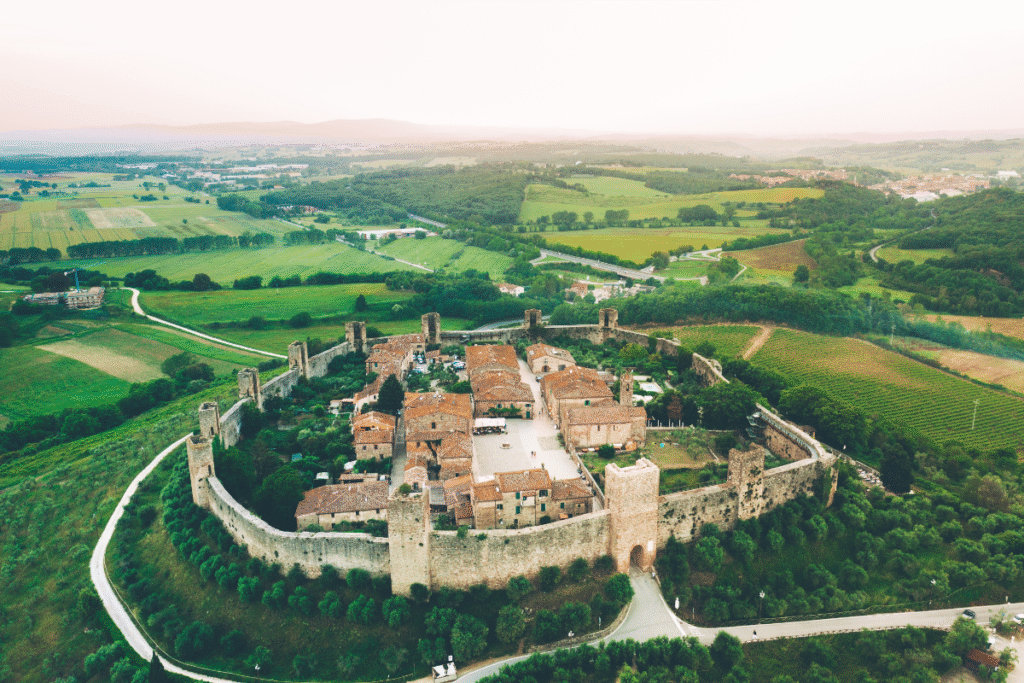 Aerial view of Castello di Monteriggioni in Tuscany, Italy, showing its perfectly preserved circular walls and medieval towers surrounded by green fields and countryside.