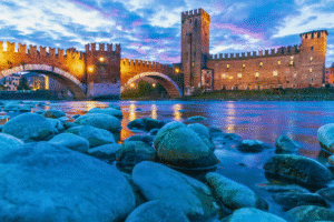 Castelvecchio and Ponte Scaligero in Verona, Italy, illuminated at twilight with reflections in the Adige River and colorful stones in the foreground.