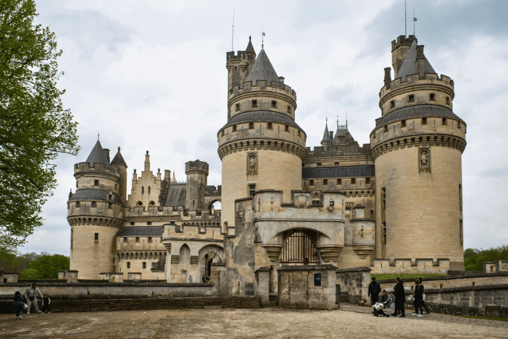 The fairytale towers and grand entrance of Château de Pierrefonds, with visitors walking near the gates on a cloudy day.