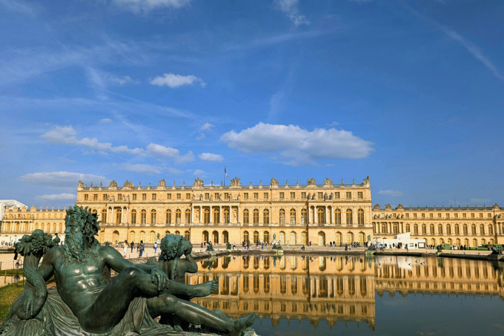 View of the Palace of Versailles reflected in the Grand Canal, with a classical garden statue in the foreground under a bright blue sky.