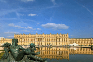 View of the Palace of Versailles reflected in the Grand Canal, with a classical garden statue in the foreground under a bright blue sky.
