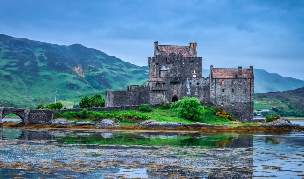 Eilean Donan Castle and arched stone bridge at sunset, surrounded by loch and green Scottish hills under dramatic clouds
