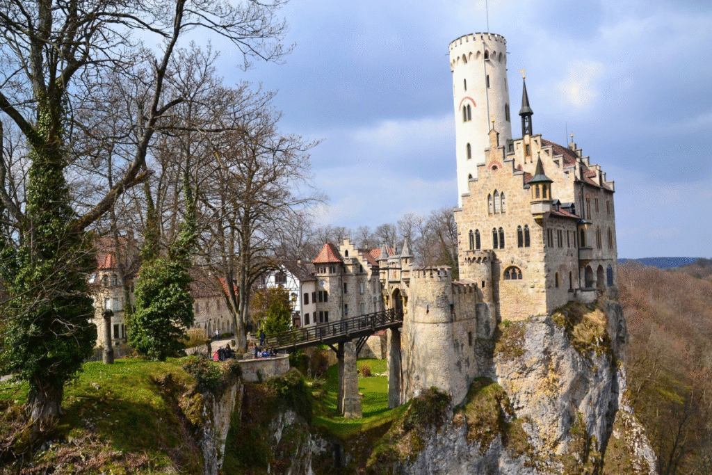 Lichtenstein Castle perched dramatically on a cliff edge, with its tall white tower, medieval-style bridge, and forested hills in the background.