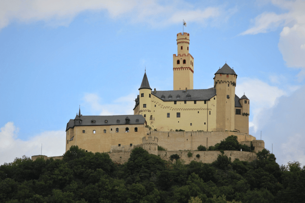Marksburg Castle perched atop a hill with its distinctive white walls and tall central tower under a blue sky, overlooking the Rhine Valley.