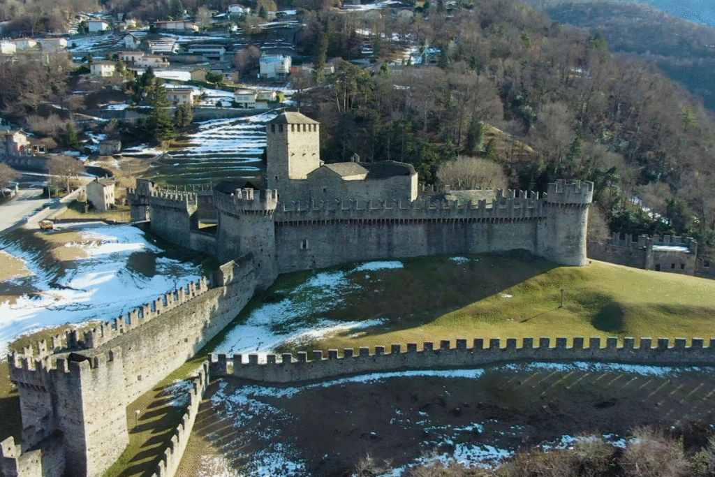 Aerial view of Montebello Castle in Switzerland, with its medieval stone walls and towers surrounded by patches of snow and hillside houses.