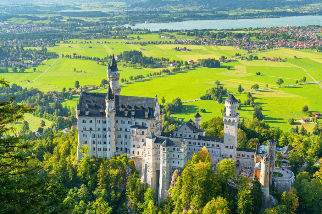 Aerial view of Neuschwanstein Castle perched above lush green meadows and a distant lake in Bavaria, Germany.