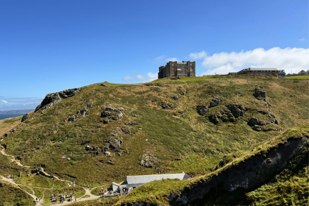 Tintagel Castle ruins perched atop rugged green cliffs in Cornwall, with visitors exploring paths below and blue sky overhead.