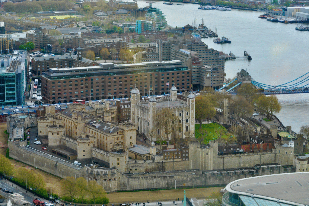 Aerial view of the Tower of London’s historic stone walls and White Tower, with the River Thames and Tower Bridge in the background.