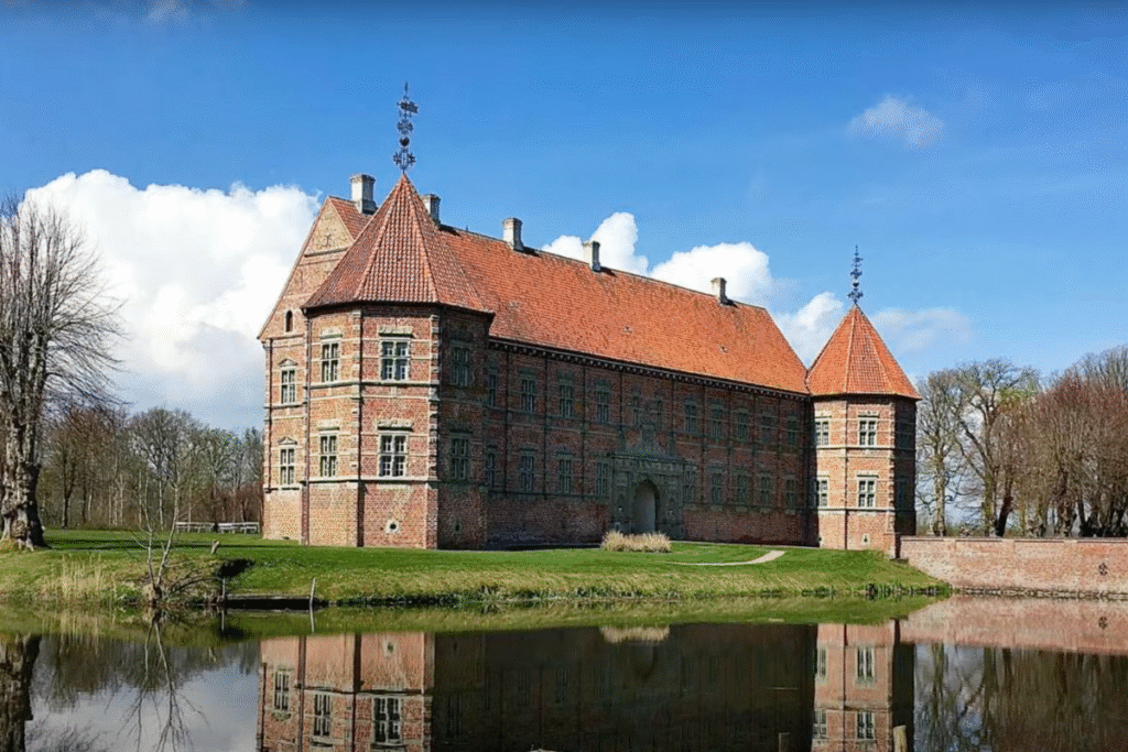 Voergaard Castle in Denmark, reflected in a still moat under a bright blue sky with red-brick walls and turrets.