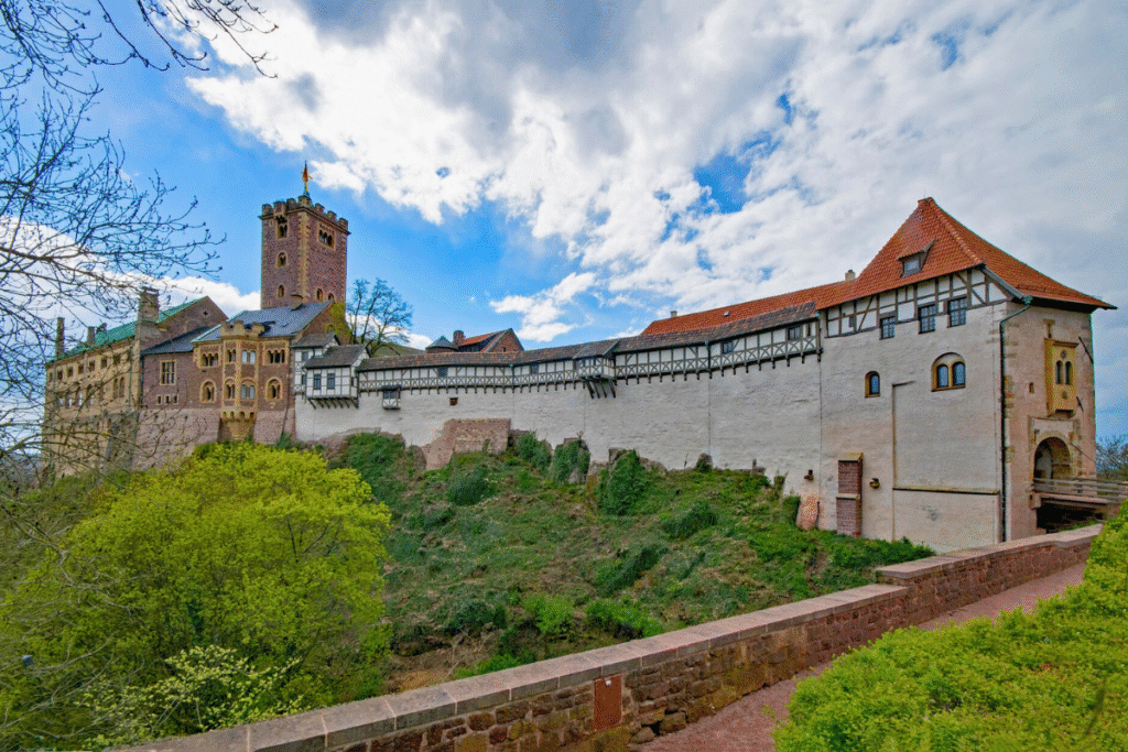 Wartburg Castle’s fortified walls and tower rise above the green hillside in Thuringia, Germany, beneath a blue sky.