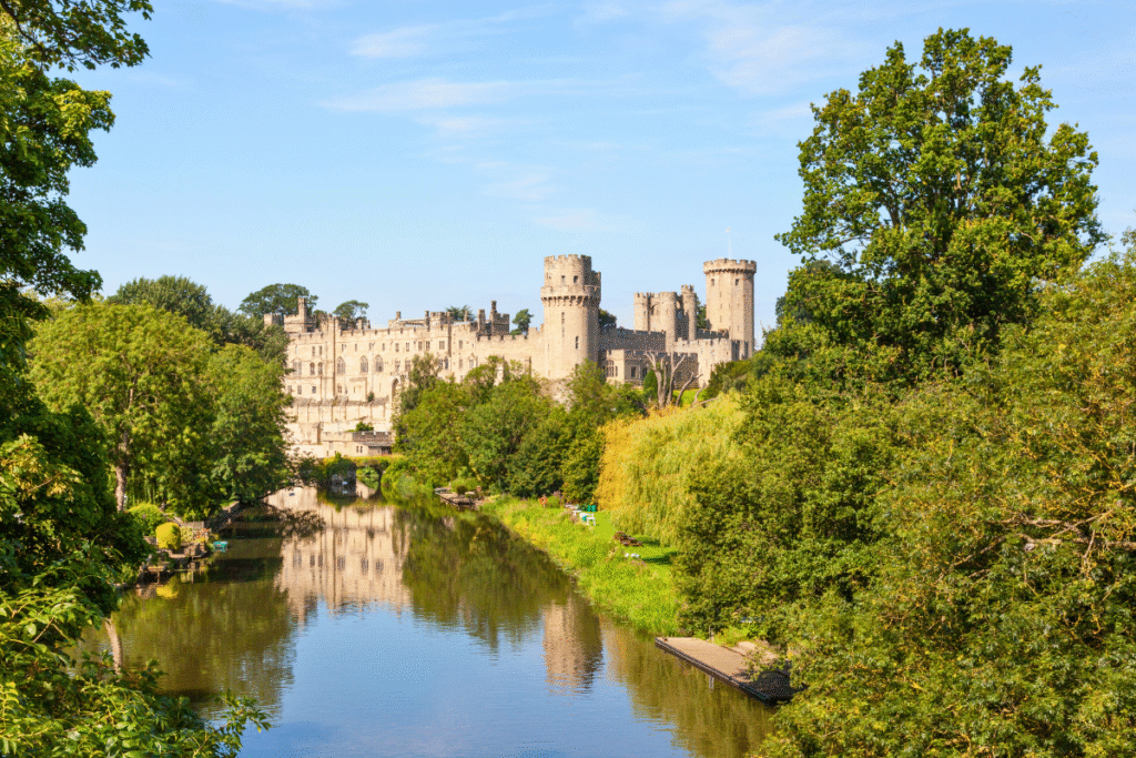Warwick Castle reflected in the calm waters of the River Avon, surrounded by lush green trees on a sunny day.