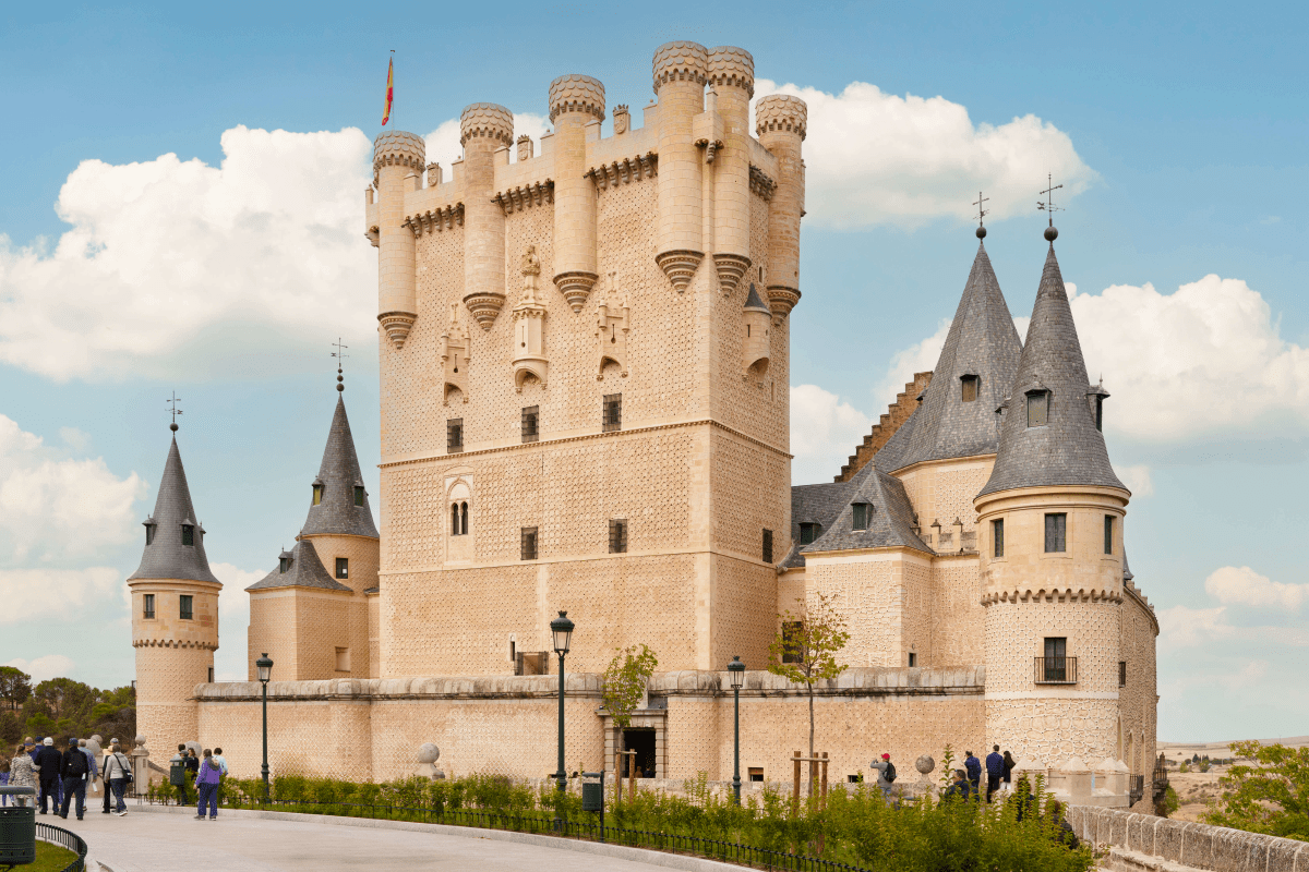 People walking by the Alcázar of Segovia castle under a blue sky.