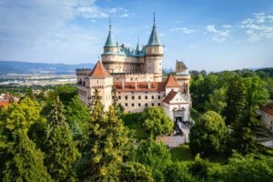 Bojnice Castle Slovakia surrounded by lush green trees and a scenic valley backdrop under a blue sky