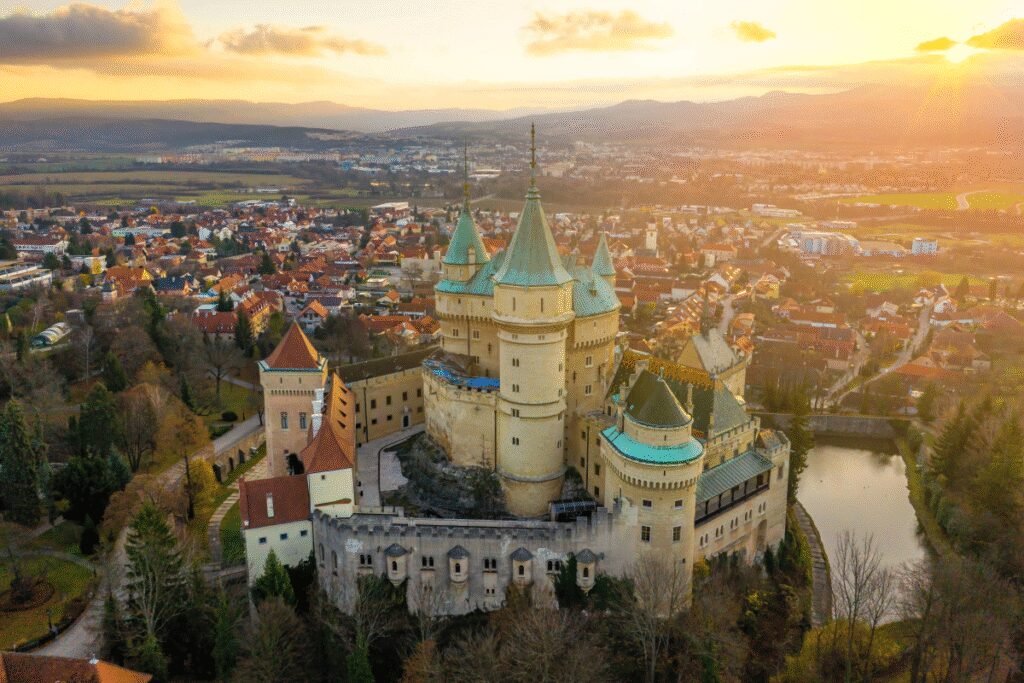 Aerial view of Bojnice Castle at sunset with town and hills in background.