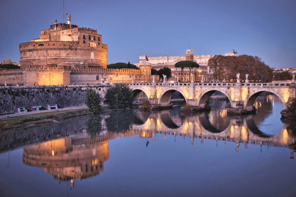 Castel Sant’Angelo illuminated at sunset beside Rome’s Tiber River and historic bridge.