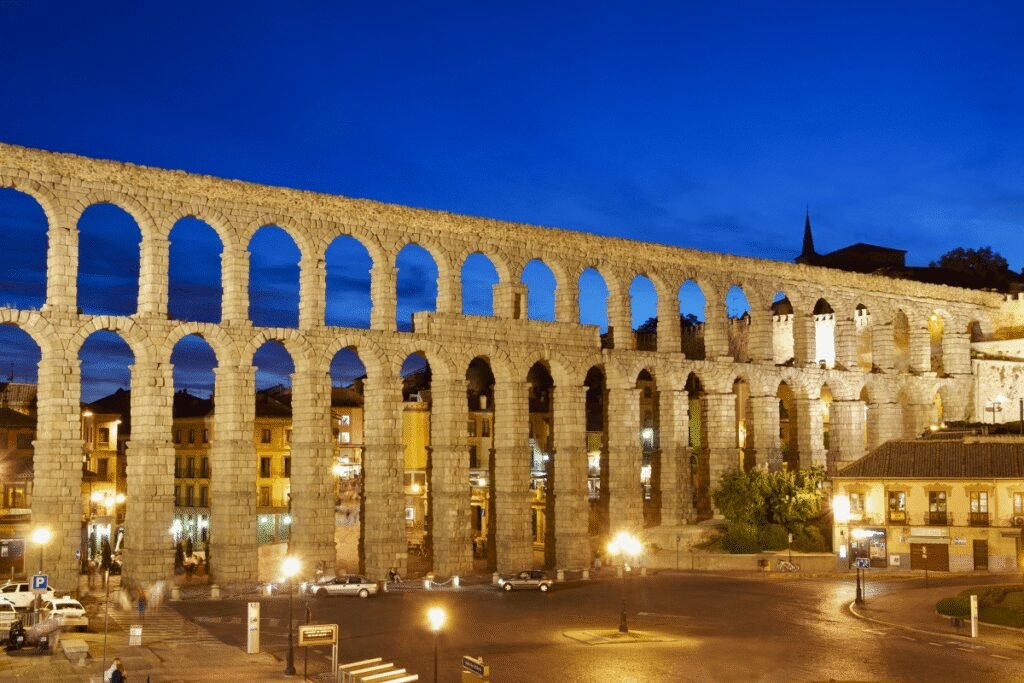 Roman Aqueduct of Segovia illuminated at dusk with arches and city lights visible.