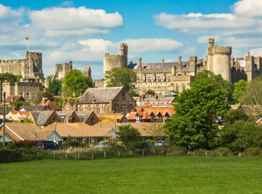 Panoramic view of Arundel Castle above rooftops and green fields under blue sky in West Sussex.